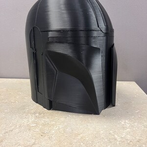 Puede incluir: Un casco negro mandaloriano, probablemente impreso en 3D, con un acabado liso y mate. El casco presenta la ic&oacute;nica visera en T y un dise&ntilde;o angular, caracter&iacute;stico de la armadura del personaje. El casco se muestra sobre una superficie de tono neutro.