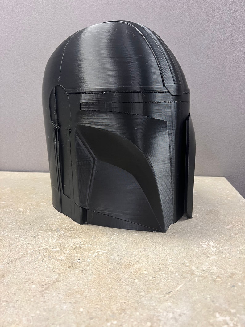 Puede incluir: Un casco negro mandaloriano, probablemente impreso en 3D, con un acabado liso y mate. El casco presenta la ic&oacute;nica visera en T y un dise&ntilde;o angular, caracter&iacute;stico de la armadura del personaje. El casco se muestra sobre una superficie de tono neutro.