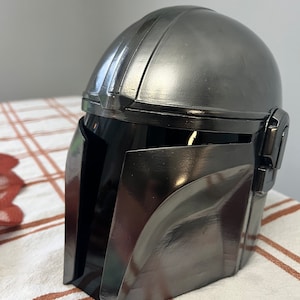 Puede incluir: Un casco gris met&aacute;lico con una visera oscura, que recuerda a un casco mandaloriano. El casco tiene una superficie lisa y reflectante con bordes definidos y una parte superior redondeada. El dise&ntilde;o presenta una visera y detalles laterales, lo que sugiere un tema futurista o de ciencia ficci&oacute;n.
