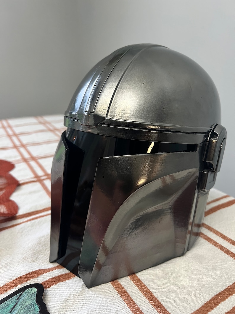 Puede incluir: Un casco gris met&aacute;lico con una visera oscura, que recuerda a un casco mandaloriano. El casco tiene una superficie lisa y reflectante con bordes definidos y una parte superior redondeada. El dise&ntilde;o presenta una visera y detalles laterales, lo que sugiere un tema futurista o de ciencia ficci&oacute;n.