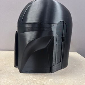 Puede incluir: Un casco negro mandaloriano, impreso en 3D con l&iacute;neas de capa visibles. El casco presenta una parte superior redondeada, una visera en &aacute;ngulo y secciones detalladas de mejillas y mand&iacute;bula. El casco se muestra sobre una superficie clara.