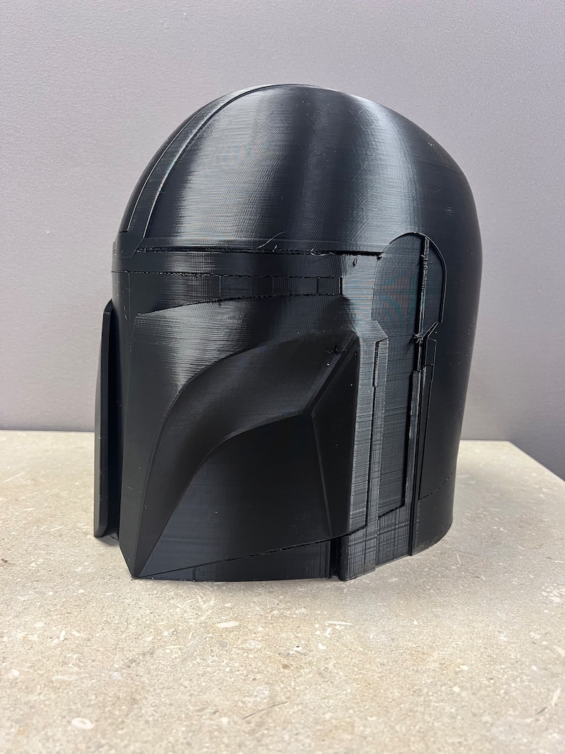 Puede incluir: Un casco negro mandaloriano, impreso en 3D con l&iacute;neas de capa visibles. El casco presenta una parte superior redondeada, una visera en &aacute;ngulo y secciones detalladas de mejillas y mand&iacute;bula. El casco se muestra sobre una superficie clara.
