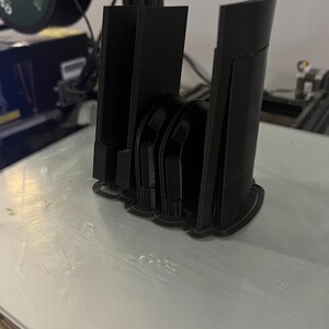 Puede incluir: Se muestra un objeto negro impreso en 3D, posiblemente un soporte para tel&eacute;fono o un dispositivo similar. El objeto tiene una serie de soportes verticales y una base curva. El art&iacute;culo est&aacute; sobre una superficie de color claro, probablemente una cama de impresora.