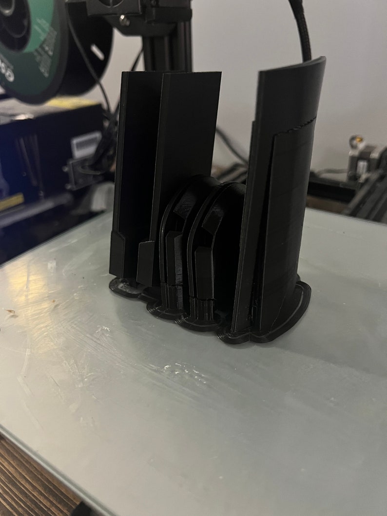 Puede incluir: Se muestra un objeto negro impreso en 3D, posiblemente un soporte para tel&eacute;fono o un dispositivo similar. El objeto tiene una serie de soportes verticales y una base curva. El art&iacute;culo est&aacute; sobre una superficie de color claro, probablemente una cama de impresora.