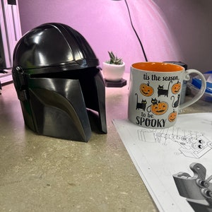 Puede incluir: Un casco negro mandaloriano con un acabado brillante est&aacute; junto a una taza de Halloween que dice "tis the season to be spooky". Tambi&eacute;n se ve una peque&ntilde;a planta suculenta en una maceta blanca.