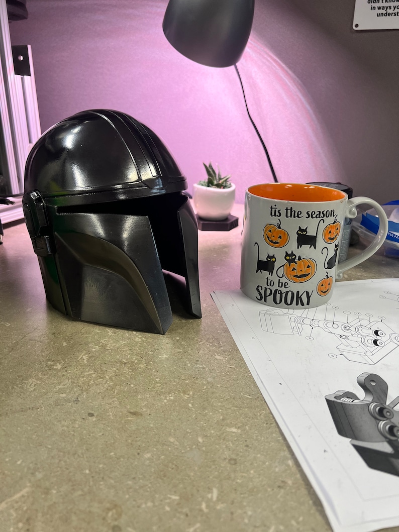 Puede incluir: Un casco negro mandaloriano con un acabado brillante est&aacute; junto a una taza de Halloween que dice "tis the season to be spooky". Tambi&eacute;n se ve una peque&ntilde;a planta suculenta en una maceta blanca.