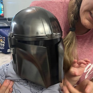 Puede incluir: Un casco Mandaloriano gris oscuro, una r&eacute;plica de la serie Star Wars. El casco tiene un acabado met&aacute;lico elegante con paneles detallados y una visera. El casco est&aacute; siendo sostenido por una persona.
