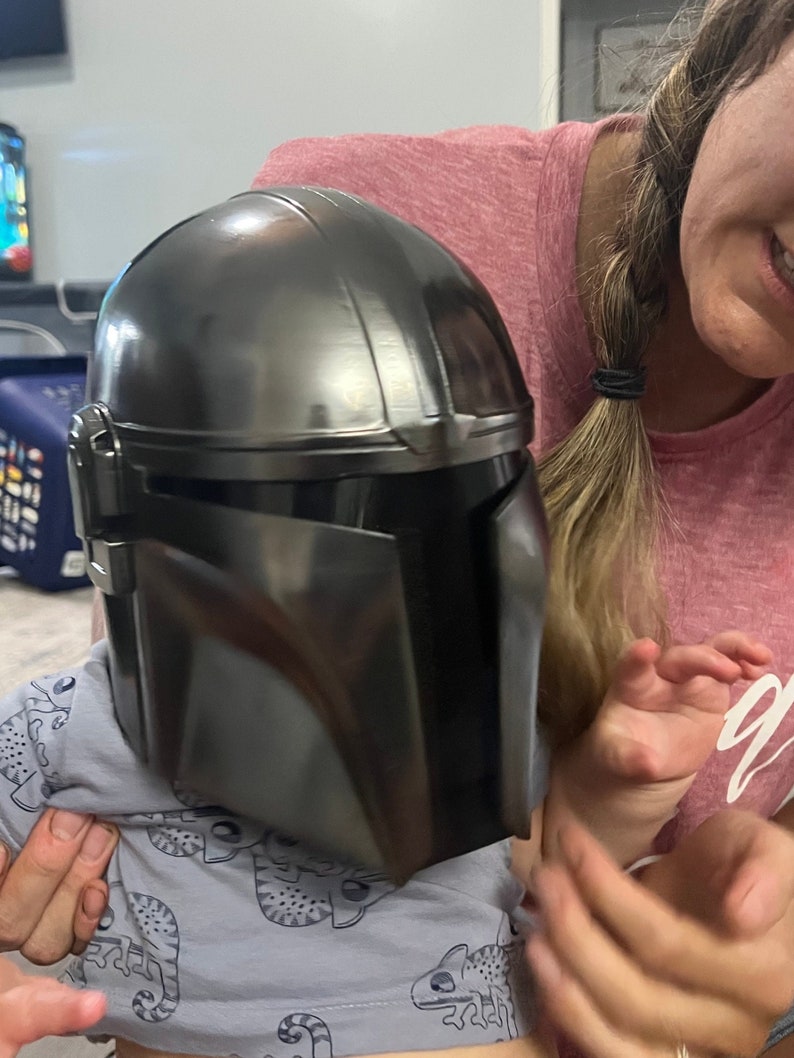 Puede incluir: Un casco Mandaloriano gris oscuro, una r&eacute;plica de la serie Star Wars. El casco tiene un acabado met&aacute;lico elegante con paneles detallados y una visera. El casco est&aacute; siendo sostenido por una persona.