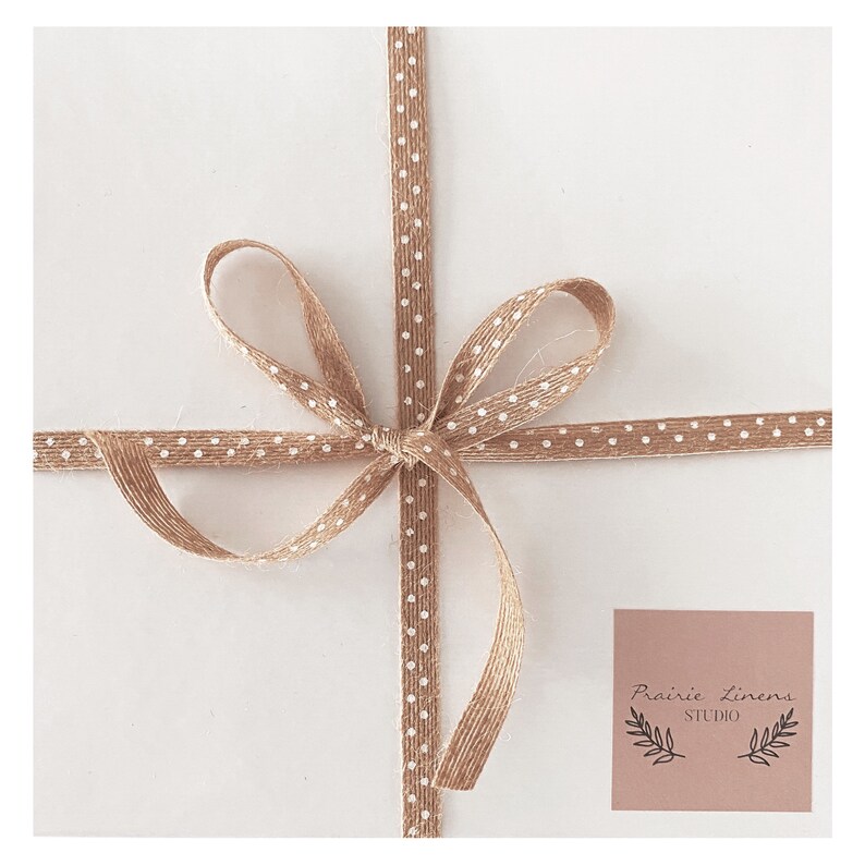 Puede incluir: Una caja de regalo blanca atada con una cinta marr&oacute;n con lunares blancos. La cinta est&aacute; atada en un lazo. La caja tiene una etiqueta marr&oacute;n con el texto "Prairie Linens Studio" en blanco.