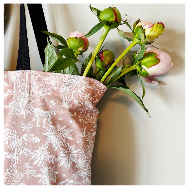 Puede incluir: Una bolsa de tela rosa y blanca con estampado tropical de monos y palmeras. La bolsa est&aacute; llena de un ramo de peon&iacute;as rosas y verdes.