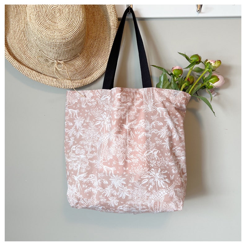 Puede incluir: Una bolsa de tela rosa con un estampado blanco de palmeras y jirafas. La bolsa tiene asas negras y est&aacute; colgada de un gancho.