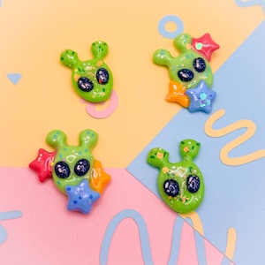 Op de afbeelding: Vier groene alien-vormige bedels met grote zwarte ogen en steraccenten in rood, oranje en blauw. De bedels hebben een glanzende afwerking en zijn verspreid op een kleurrijke achtergrond van roze, geel en blauw.