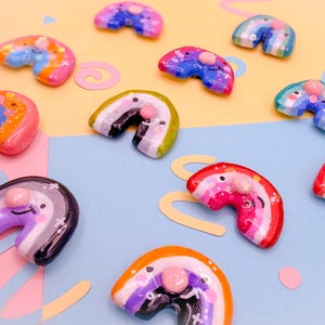 May include: A collection of colorful, miniature rainbow-shaped charms with a small, round, pink bead at the center. The charms are painted in various colors, including pink, blue, orange, and purple. Some have glitter accents. The background is a mix of pink and blue.
