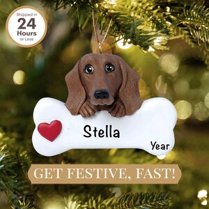 May include: A personalized Christmas ornament featuring a brown dachshund dog with the name "Stella" on a white bone-shaped plaque. The ornament has a red heart and the text "Year" on the plaque.