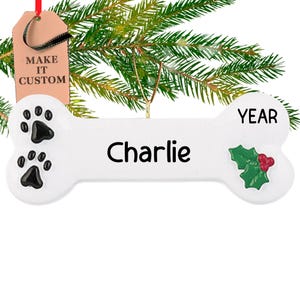 Op de afbeelding: Witte kerstversiering in de vorm van een bot met zwarte poot afdrukken, de naam "Charlie" en het woord "YEAR" erop. De versiering heeft een groene hulsttak met rode bessen.