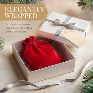 May include: A white gift box with a silver ribbon and a blank tag is open to reveal a red velvet drawstring bag. The text "Elegantly Wrapped" is on a gold background above the box. The text "Our Optional Velvet Bag & Luxe Box Make Gifting a Delight" is below the box.