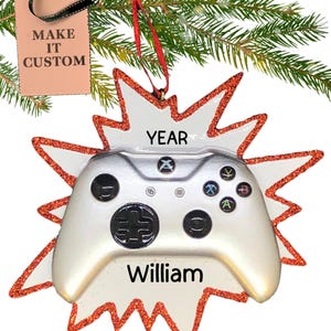 Peut inclure: Ornement de Noël en forme de manette de jeu vidéo blanche et argentée avec des paillettes rouges sur les bords. L'ornement porte les inscriptions "YEAR" et "William".