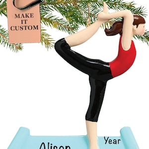 Op de afbeelding: Een gepersonaliseerde kerstversiering met een vrouw in een rood tanktop en zwarte leggings die een yogapose uitvoert op een blauwe yogamat. De mat heeft de naam "Alison" en het woord "Jaar" erop geschreven.