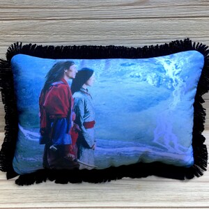 May include: A black fringed pillow with a blue and white image of two people standing in front of a blue and white water scene.