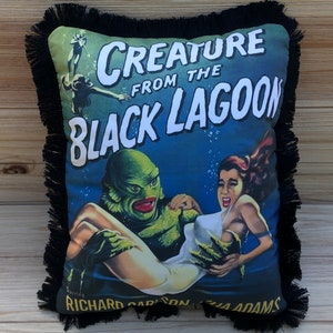 May include: A black and green pillow with a fringe border. The pillow features a vintage movie poster design for the film "Creature from the Black Lagoon" with the text "Creature from the Black Lagoon" in white letters. The poster features a green creature holding a woman in a white swimsuit.