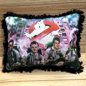 May include: A black pillow with a fringe border features a colorful print of the Ghostbusters characters in their iconic jumpsuits, driving their car, with the Ghostbusters logo and a ghost in the background.