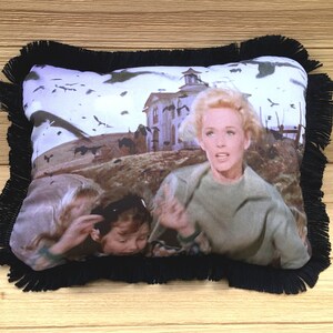 May include: A black and white pillow with a fringe border features a scene from the movie "The Birds" with Tippi Hedren and two children running from a flock of birds.