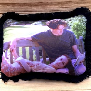 May include: Rectangular pillow with a printed image of two people on a park bench, with a black fringe border. The image shows a man and a woman, with the woman lying down and the man sitting.