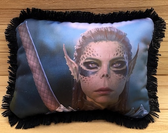 Puede incluir: Una almohada decorativa con un borde de flecos negros presenta una imagen de primer plano de una mujer con ojos azules, piel verde y una oreja puntiaguda. Lleva una armadura de cuero oscura y tiene una espada en el fondo.