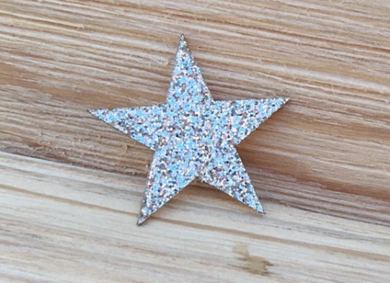 May include: A silver glitter star with five points. The star is on a light brown wooden surface.