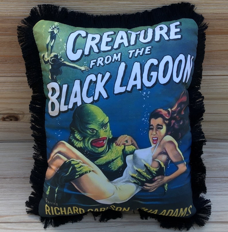 May include: A black and green pillow with a fringe border. The pillow features a vintage movie poster design for the film "Creature from the Black Lagoon" with the text "Creature from the Black Lagoon" in white letters. The poster features a green creature holding a woman in a white swimsuit.