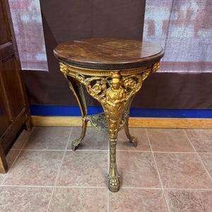 Puede incluir: Una mesa auxiliar de hierro fundido pintada de oro con una tapa redonda de madera y patas ornamentadas. La mesa presenta un diseño decorativo con una figura en el centro.