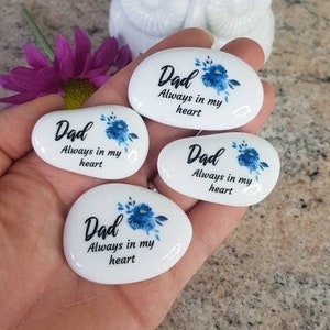 May include: Four white oval-shaped stones with blue floral designs and the text "Dad Always in my heart" on each stone.