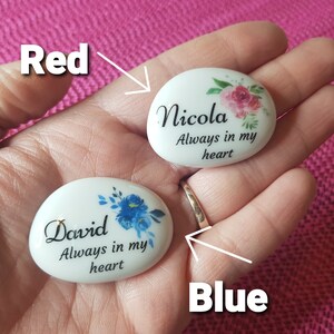 May include: Two white, oval-shaped memorial stones with floral designs and the text "Nicola Always in my heart" and "David Always in my heart". The Nicola stone has pink flowers, and the David stone has blue flowers.