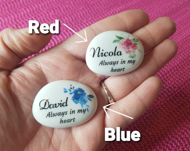 May include: Two white, oval-shaped memorial stones with floral designs and the text "Nicola Always in my heart" and "David Always in my heart". The Nicola stone has pink flowers, and the David stone has blue flowers.