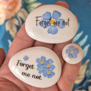 May include: Three white, oval stones featuring blue forget-me-not flower designs and the words "Forget me not" in black script. The stones are different sizes, including a small, round one.