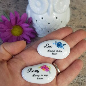 May include: Two small, white, oval-shaped stones with floral designs and names, "Leo" and "Roxy," are held in a hand. The text "Always in my heart" is below the names. A white owl-shaped oil warmer and a purple flower are in the background.