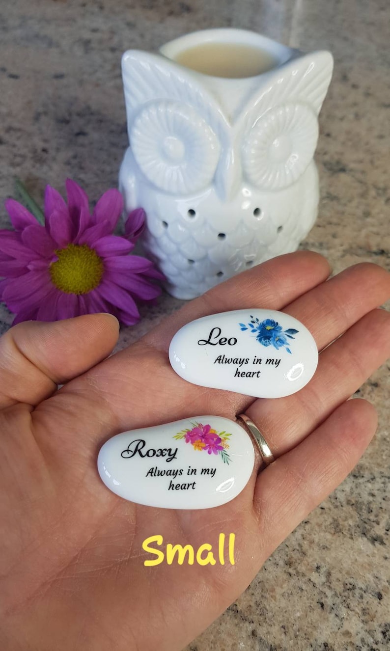 May include: Two small, white, oval-shaped stones with floral designs and names, "Leo" and "Roxy," are held in a hand. The text "Always in my heart" is below the names. A white owl-shaped oil warmer and a purple flower are in the background.