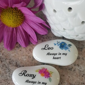 May include: Two white, oval-shaped ceramic stones with floral designs and the names "Leo" and "Roxy" printed on them. The text "Always in my heart" is below each name. A white owl-shaped ceramic diffuser and a pink flower are in the background.