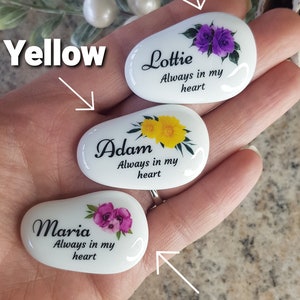 May include: Three white, oval-shaped stones with floral designs and names. One stone has purple flowers and the name "Lottie", another has yellow flowers and the name "Adam", and the last has pink flowers and the name "Maria". Each stone has the phrase "Always in my heart".