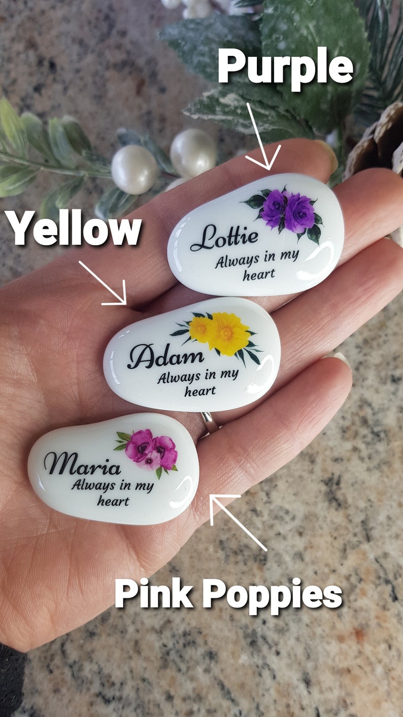 May include: Three white, oval-shaped stones with floral designs and names. One stone has purple flowers and the name "Lottie", another has yellow flowers and the name "Adam", and the last has pink flowers and the name "Maria". Each stone has the phrase "Always in my heart".