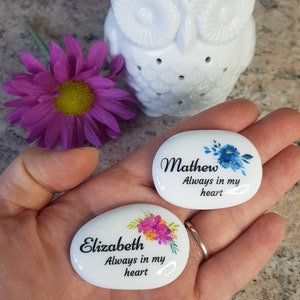 May include: Two white, oval-shaped memorial stones with floral designs and names, "Mathew" and "Elizabeth," with the text "Always in my heart." A white owl-shaped essential oil diffuser and a purple flower are also in the image.