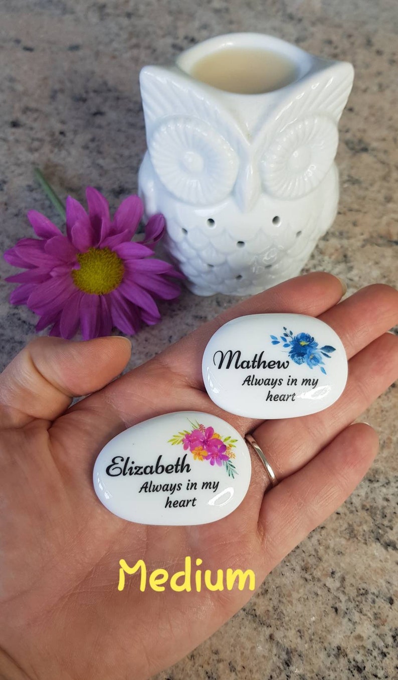 May include: Two white, oval-shaped memorial stones with floral designs and names, "Mathew" and "Elizabeth," with the text "Always in my heart." A white owl-shaped essential oil diffuser and a purple flower are also in the image.
