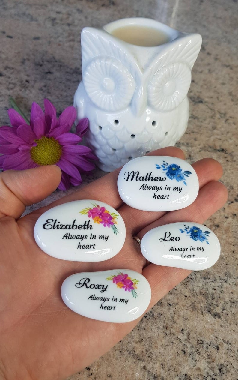 May include: White, oval-shaped memorial stones with names and floral designs. Each stone has a name and the phrase "Always in my heart." A white owl-shaped essential oil diffuser and a purple flower are in the background.