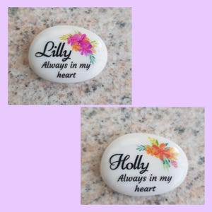 May include: Two white, oval-shaped stones with floral designs and text. One stone says "Lilly Always in my heart" with pink and yellow flowers. The other says "Holly Always in my heart" with orange and yellow flowers.
