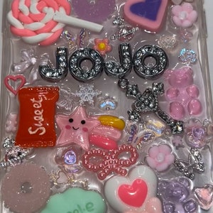 Peut inclure: Une coque de téléphone transparente décorée de breloques colorées, dont une étoile rose, une barre chocolatée rouge avec le mot "Sweet" dessus, une sucette rose, un charme argenté avec le mot "Jojo" dessus et un nuage vert avec le mot "Angel.e" dessus.