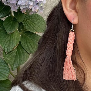 May include: A peach-coloured macrame tassel earring is shown close-up. The earring hangs from a silver hook and has a braided design leading to a fringed tassel. The background features green leaves and a light purple flower.