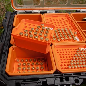 May include: An open black and orange storage case with multiple compartments filled with brass ammunition. The compartments are labelled with the ammunition type. The case is on a grassy surface.