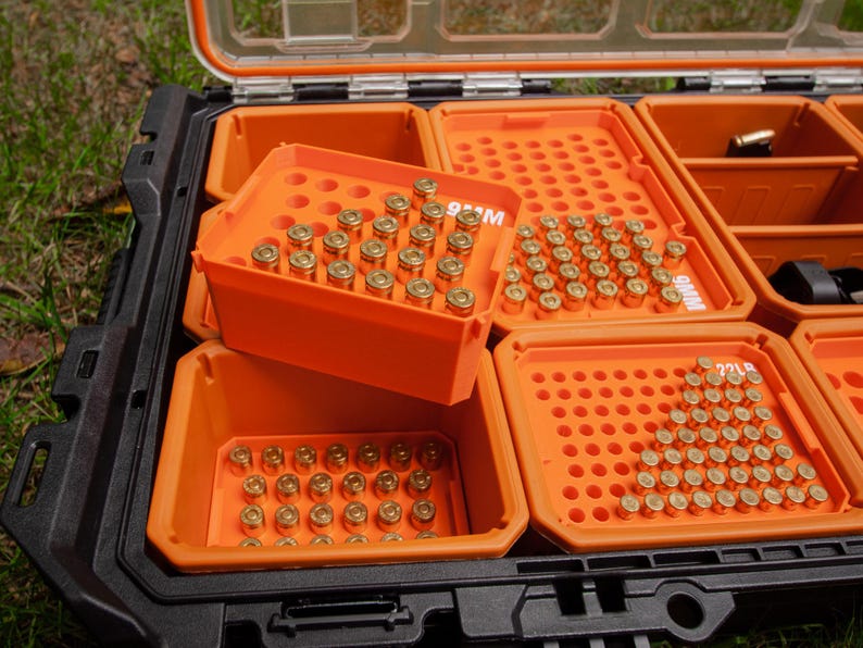 May include: An open black and orange storage case with multiple compartments filled with brass ammunition. The compartments are labelled with the ammunition type. The case is on a grassy surface.