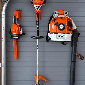 May include: Three Stihl power tools mounted on a grey wall: a chainsaw on the left, a string trimmer labelled FS 91 R in the centre, and a backpack leaf blower labelled BR 600 on the right. All tools feature the signature orange and white colour scheme.