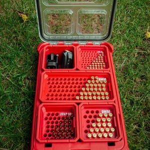 Puede incluir: Caja de almacenamiento de plástico roja abierta con tapa transparente, que revela compartimentos llenos de munición. La caja está etiquetada con "Milwaukee" y varias marcas de calibre, incluyendo "9MM" y "223/F 56".